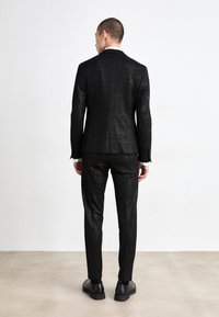 Fekete öltöny, finom fényes textúrával, slim-fit fazonban, és egyetlen hátsó szellőzővel. Fekete cipővel párosítva, hátulról nézve.