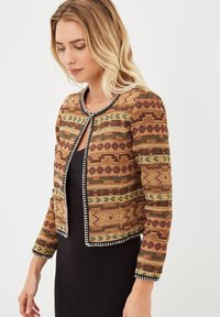 Mujer vistiendo una chaqueta tejida de colores marrón, verde y rojo con ribetes negros y blancos sobre un vestido negro, mirando ligeramente hacia abajo.