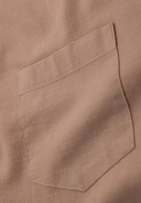 Tissu marron clair avec une texture visible, comportant une poche plaquée sur le côté gauche. Design simple avec des coutures nettes et des bords droits.