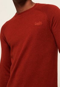 Maglione rosso lavorato a maglia con scollatura rotonda, polsini a coste e dettaglio del logo ricamato sul petto. Texture morbida, design aderente e maniche lunghe.
