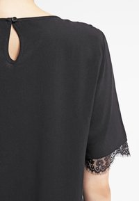 Svart kortärmad topp med spetsdetaljer vid ärmarna och en nyckelhålsöppning bak, tillverkad av ett mjukt, lätt tyg.