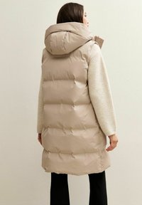 Gilet imbottito beige con cappuccio, caratterizzato da sezioni trapuntate orizzontali, colletto alto e una vestibilità rilassata. Indossato sopra un maglione leggero e pantaloni neri.