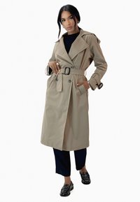 Manteau beige avec ceinture, design double boutonnage, grand col et poches latérales. Porté sur un haut noir et un pantalon bleu marine.