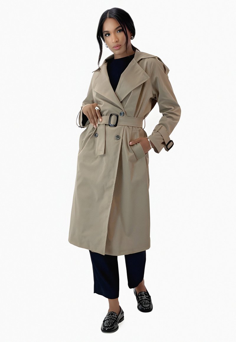 Manteau beige avec ceinture, design double boutonnage, grand col et poches latérales. Porté sur un haut noir et un pantalon bleu marine.