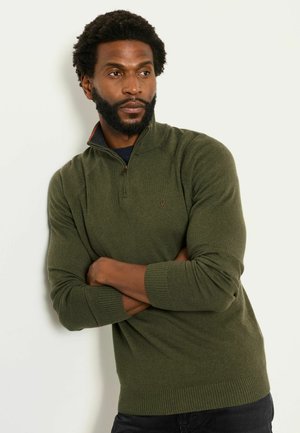 Mann mit Bart und lockigem Haar, der einen olivgrünen Half-Zip-Pullover und schwarze Hosen trägt, steht mit verschränkten Armen vor einem schlichten Hintergrund.