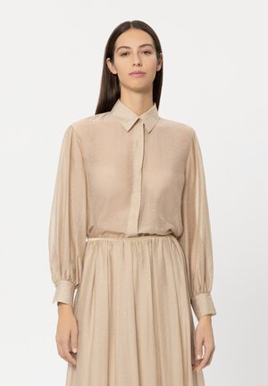 Beige blouse with a button-down collar and long, billowy sleeves, featuring a subtle texture and sparkly accents. Paired with a matching skirt.
