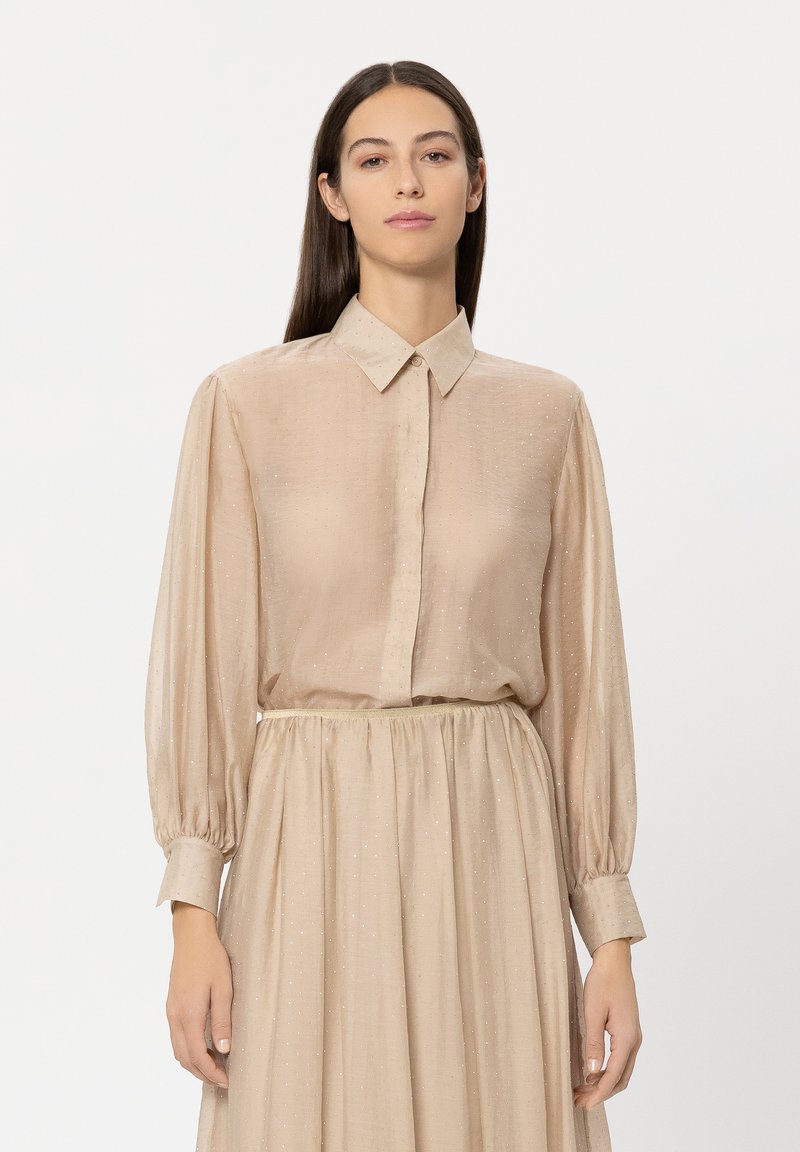 Beige blouse with a button-down collar and long, billowy sleeves, featuring a subtle texture and sparkly accents. Paired with a matching skirt.