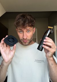 Mies, jolla on vaalea parta ja kiharat hiukset, pitää toisessa kädessä mustaa sähköistä hiustenleikkuukonetta ja toisessa sen latausalustaa.
