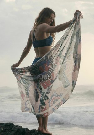 Woman in blue swimwear standing barefoot on rocks by the sea, holding a large floral patterned sheer scarf spread behind her.