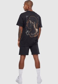 Sort bomulds t-shirt med et stort grafisk print på bagsiden, der viser et portræt og designdetaljer. Parret med sorte shorts og hvide sneakers.