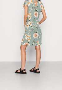 Robe florale verte à manches courtes et dos en keyhole, ornée de grandes fleurs orange et blanches, accompagnée de sandales noires à lanières.