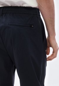 Κοντινό πλάνο ενός ατόμου που φοράει μαύρο παντελόνι με ελαστική μέση και φερμουάρ σε πίσω τσέπη, το χ έρι του χαλαρό στο πλάι.