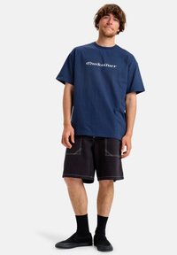 Námořnická modrá bavlněná tričko s krátkým rukávem a bílým logem "Quiksilver", spárované s černými šortkami s kontrastním švem a kapsami.