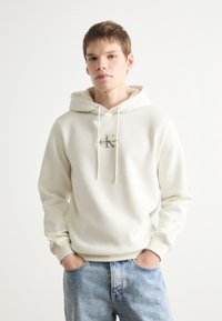 Sudadera con capucha de color crema claro, fabricada en un tejido suave, con cordones y un logo en el frontal. Combinada con jeans de mezclilla azul desgastados. Diseño simple y casual.