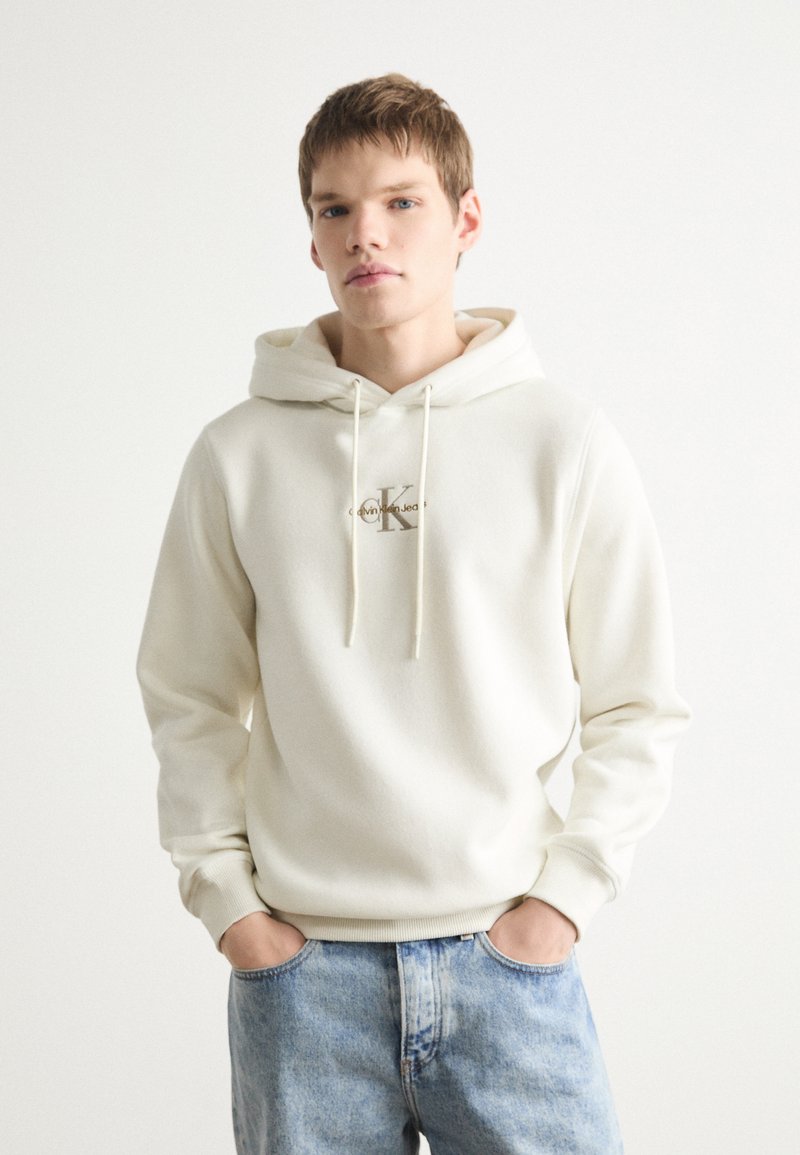 Sudadera con capucha de color crema claro, fabricada en un tejido suave, con cordones y un logo en el frontal. Combinada con jeans de mezclilla azul desgastados. Diseño simple y casual.