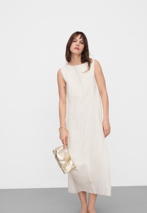 Frau mit mittelbraunen Haaren, die ein ärmelloses weißes Midikleid trägt und eine goldene Clutch vor einem schlichten grau-weißen Hintergrund hält.