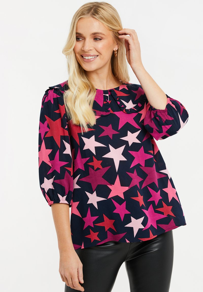 Farbige Bluse mit einem Sternenmuster in Pink, Rot und Marineblau. Sie hat einen Rüschenausschnitt und dreiviertellange Puffärmel.