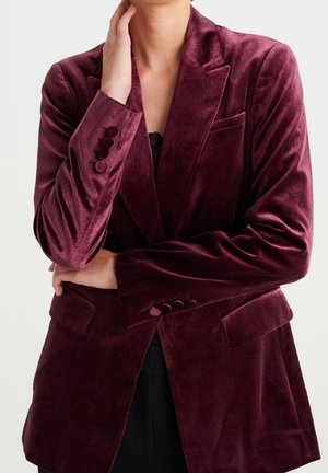 Vrouw draagt een zachte bordeauxrode fluwelen blazer met knoopdetails, waarvan één arm gekruist is en de andere hand haar hals zachtjes aanraakt.
