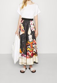 Biała bluzka z krótkim rękawem w połączeniu z czarną, malarską długą spódnicą z intensywnymi kolorami i abstrakcyjnymi wzorami. Modelka trzyma białą torbę, ma na sobie sandały.