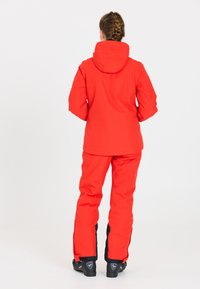 Rote Skijacke mit Kapuze und wasserdichtem Material, kombiniert mit passenden roten Hosen mit schwarzen Akzenten und Schneeschuhen.
