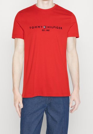 Homme portant un T-shirt rouge à manches courtes Tommy Hilfiger avec un logo et le texte « EST. 1985 », assorti à un jean bleu, sur un fond uni.