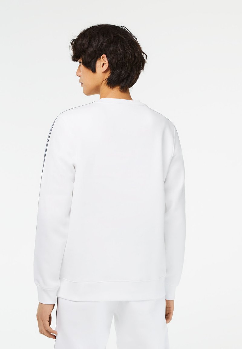 Sweatshirt blanc à manches longues, col rond et poignets côtelés. Présente une texture lisse et de subtils détails de couture le long des épaules.