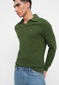 Zöld bordás pulóver v-nyakkal és széles gallérral. Kicsi logóval a mellkason. Kék farmernadrággal viselve.