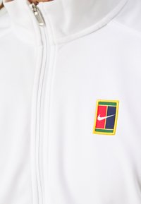 Bílá bunda na zip s vícebarevným obdélníkovým logem Nike na levé straně hrudi, obsahující červené, zelené, žluté a tmavě modré části.