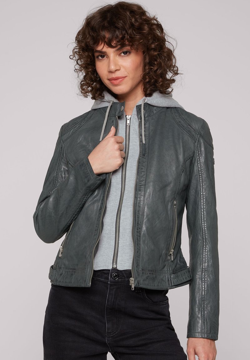 Dunkelgrüne Lederjacke mit taillierter Passform, Reißverschluss und Schulternähten. Über einem grauen Hoodie getragen, kombiniert mit schwarzen Hosen.