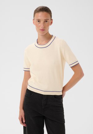 Femme aux cheveux bruns vêtue d'un pull crème à manches courtes avec un bord bleu et un jean noir, debout, une main dans le dos.