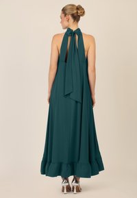 Teal halterneck kjole med en svævende silhuet, der har en bølget kant og en stor sløjfe til lukning ved halsen, vist bagfra.