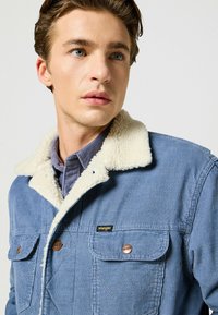 Blå manchesterjacka med en vit fuskpälskrage, bröstfickor och kontrasterande bruna knappar. Wrangler-logotyp på vänster ficka.