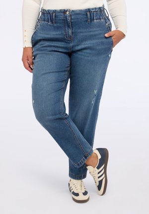 Osoba ubrana w niebieskie dżinsy z elastycznym pasem z lekkimi przetarciami, białą koszulę z długim rękawem i granatowo-białe trampki, stojąca jedną stopą przekroczoną nad drugą.