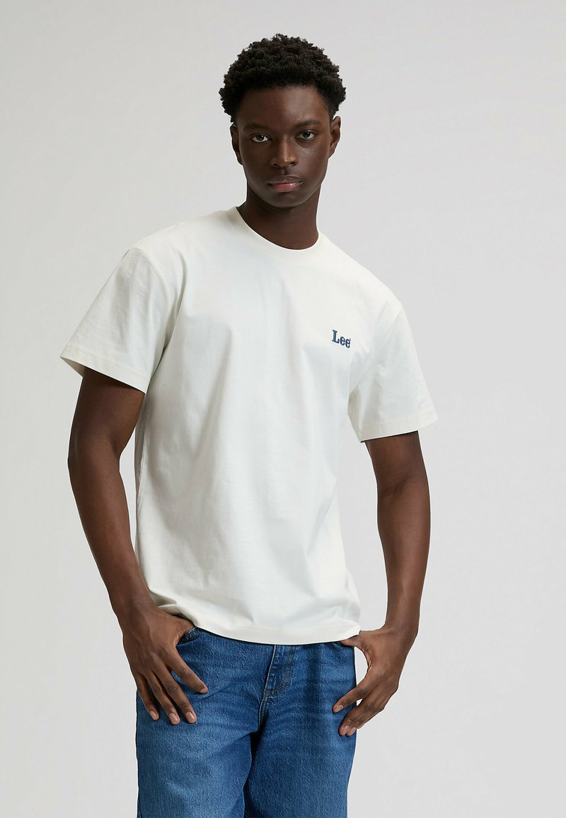 Biały bawełniany T-shirt z krótkimi rękawami i okrągłym dekoltem, z małym niebieskim logo "Lee" na piersi. Noszony z niebieskimi dżinsami.