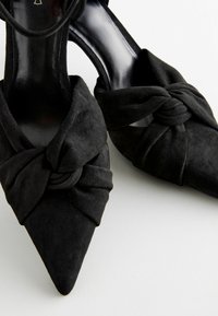 Mustat mokkasiinit, joissa on kohti suuntautuva kärki ja solmittu yksityiskohta edessä, sisältä kiiltävän mustat. Elegantti ja jämäkkä muotoilu.