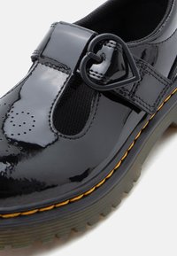 Chaussure Mary Jane en cuir verni noir avec boucle en forme de cœur, détail perforé en forme de cœur et semelle épaisse à rainures avec couture jaune.