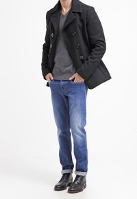 Czarny dwurzędowy płaszcz typu peacoat na szarym swetrze z dekoltem w serek, zestawiony z niebieskimi dżinsami i czarnymi skórzanymi butami.