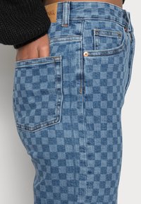 Pantalones vaqueros de mezclilla azul con un patrón de tablero de ajedrez, con un bolsillo frontal, herrajes de tono cobre y costuras de contraste. Vista parcial de la cintura.