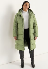 Kvinde iført en lang lysegrøn polstret frakke med hætte, hvid turtleneck, sorte bukser og sorte knæhøje støvler, der står op ad en hvid væg.