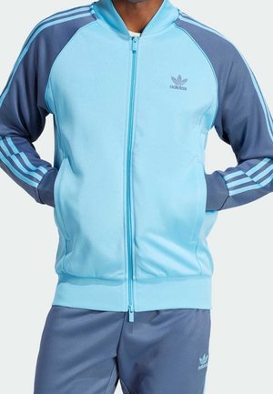 Homme portant une veste de survêtement Adidas bleu clair et bleu foncé avec un pantalon assorti, debout les mains dans les poches.