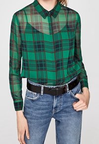 Camisa de cuadros verdes con textura transparente, con mangas largas y cuello. Combinada con jeans azul de cintura alta y un cinturón negro.