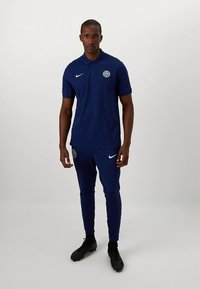 Marineblauw Nike poloshirt met kraag en logo, gecombineerd met bijpassende taps toelopende trainingsbroek. Model draagt zwarte sportschoenen.