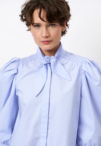 Light blue shirt with a high collar, bow tie detail, and smocked fabric on the shoulders. Puff sleeves and smooth texture.