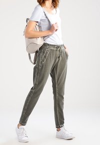 Olive zelené joggingové kalhoty vyrobené z měkké látky, s elastickým pasem, šňůrkou, bočními kapsami a zúženými kotníky. Kombinováno s lehkým topem a teniskami.