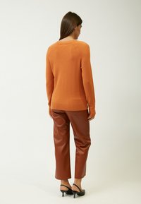 Maglione arancione lavorato a maglia con orlo e polsi a coste, abbinato a pantaloni in finta pelle marrone. La texture sembra morbida e liscia.