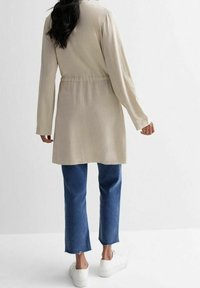 Cardigan largo beige con cinturón en la cintura, aberturas en la parte trasera y mangas largas, combinado con vaqueros azules recortados y zapatillas blancas.