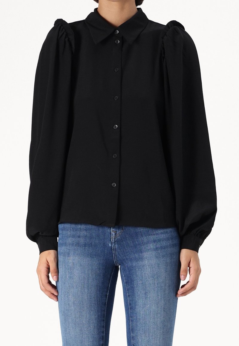 Femme portant un chemisier noir à manches longues avec des épaules bouffantes et une patte de boutonnage devant, associé à un jean bleu en denim.