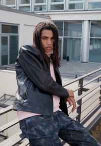 Black leather jacket, pink sweatshirt, and dark camouflage pants. The model poses with a relaxed posture against a modern urban background.