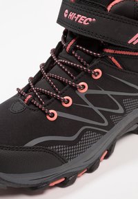 Scarponcino nero da trekking con accenti rosa. Presenta un tessuto traspirante, suola in gomma spessa, presa testurizzata e sovrapposizioni a motivi contrastanti.