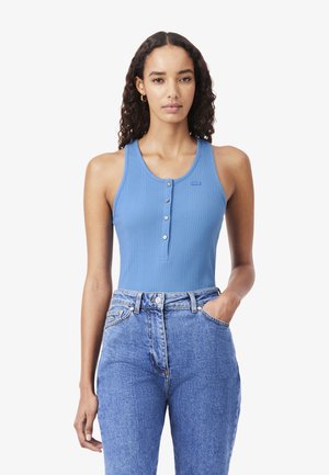 Donna con capelli ricci che indossa un top blu senza maniche a coste con bottoni e jeans blu a vita alta con la mano in tasca.